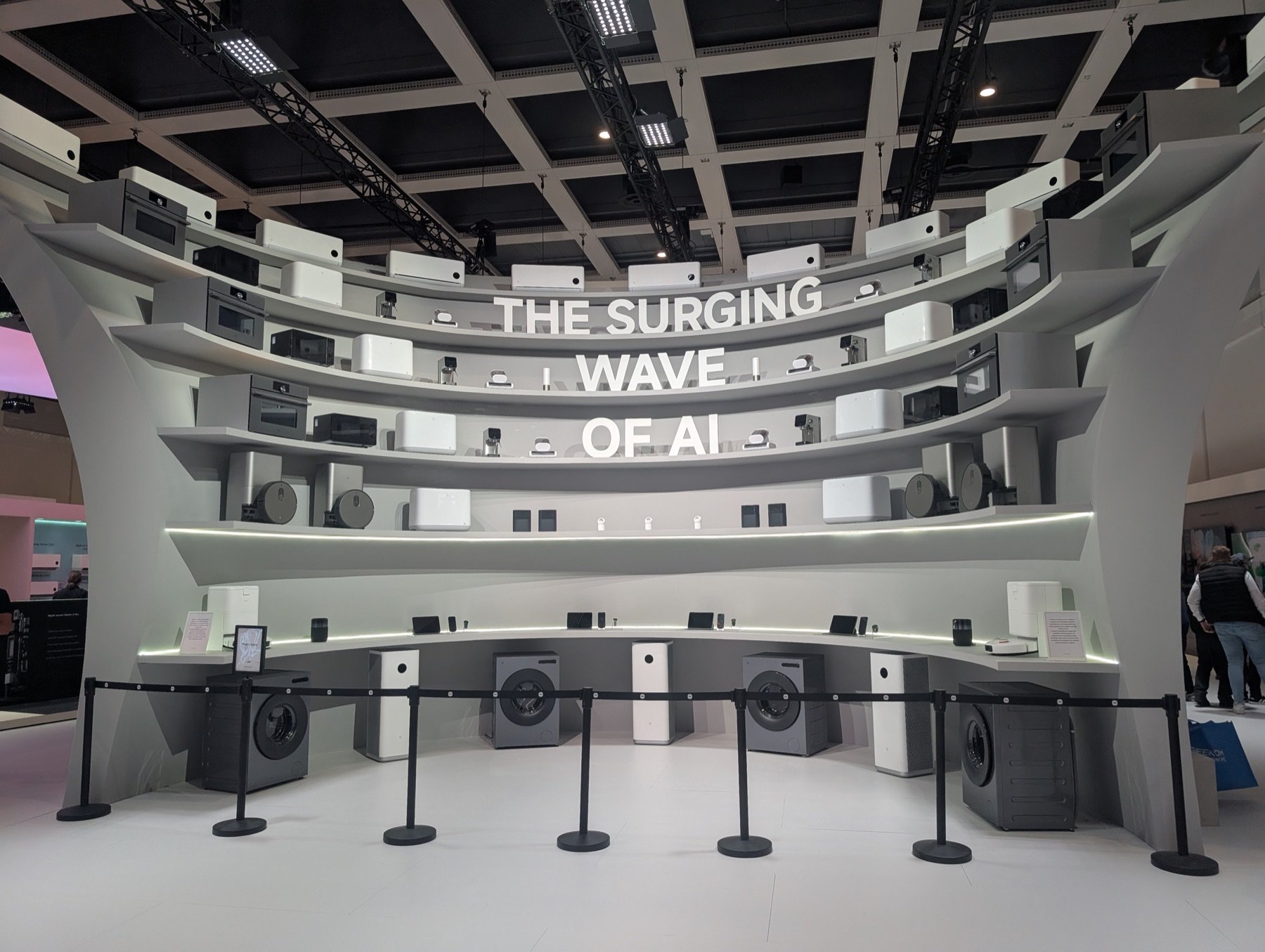 Geräte auf einer geschwungenen weißen Wand unter dem Schriftzug THE SURGING WAVE OF AI